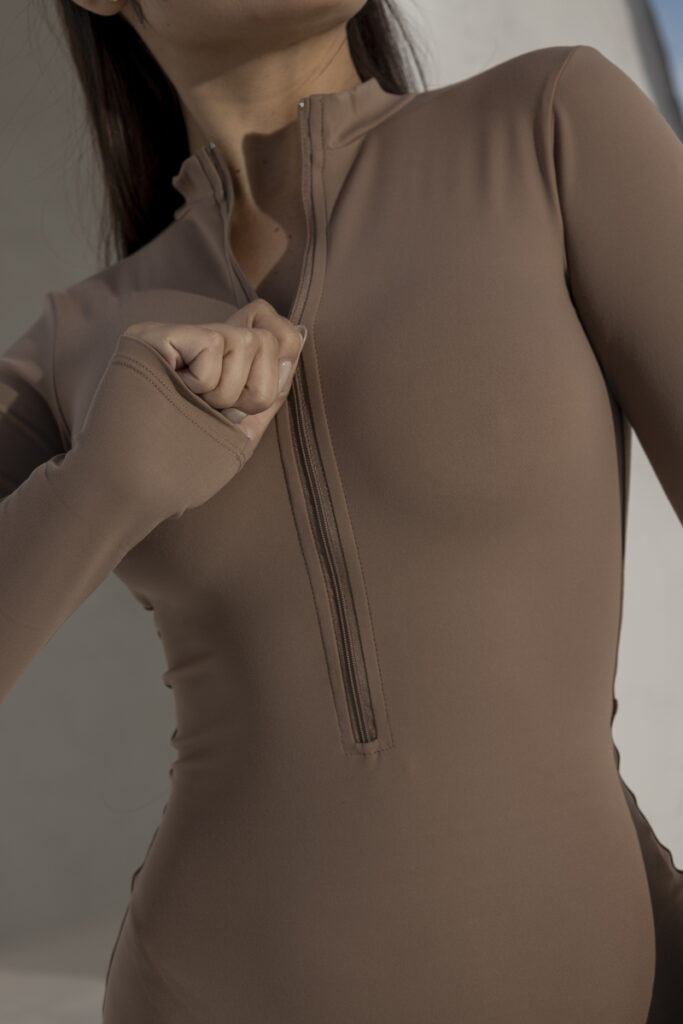 Woman wearing a fitted brown long-sleeve bodysuit with a half-zipper, showcasing a stylish and modern design against a neutral background.